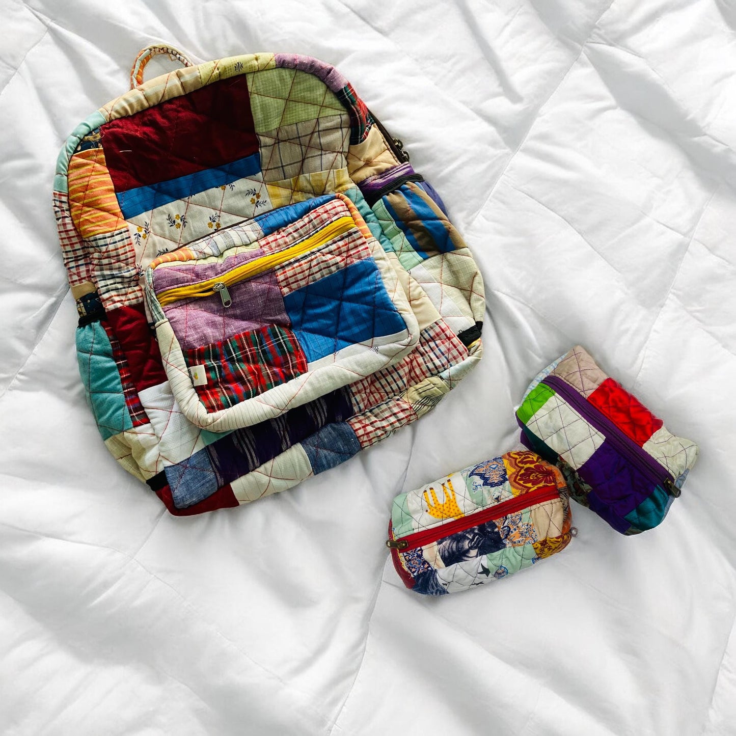 Colorful backpack and quilted pouches resting on a white duvet. Made using patchwork of various upcycled fabrics and vibrant zippers, the set is a cheerful, sustainable alternative to plastic bags. Great for school, travel, or gifting. Handcrafted by artisans, it suits toddlers, teens, and women. Perfect for baby shower gifts or as a unique accessory set for minimalist and conscious living.