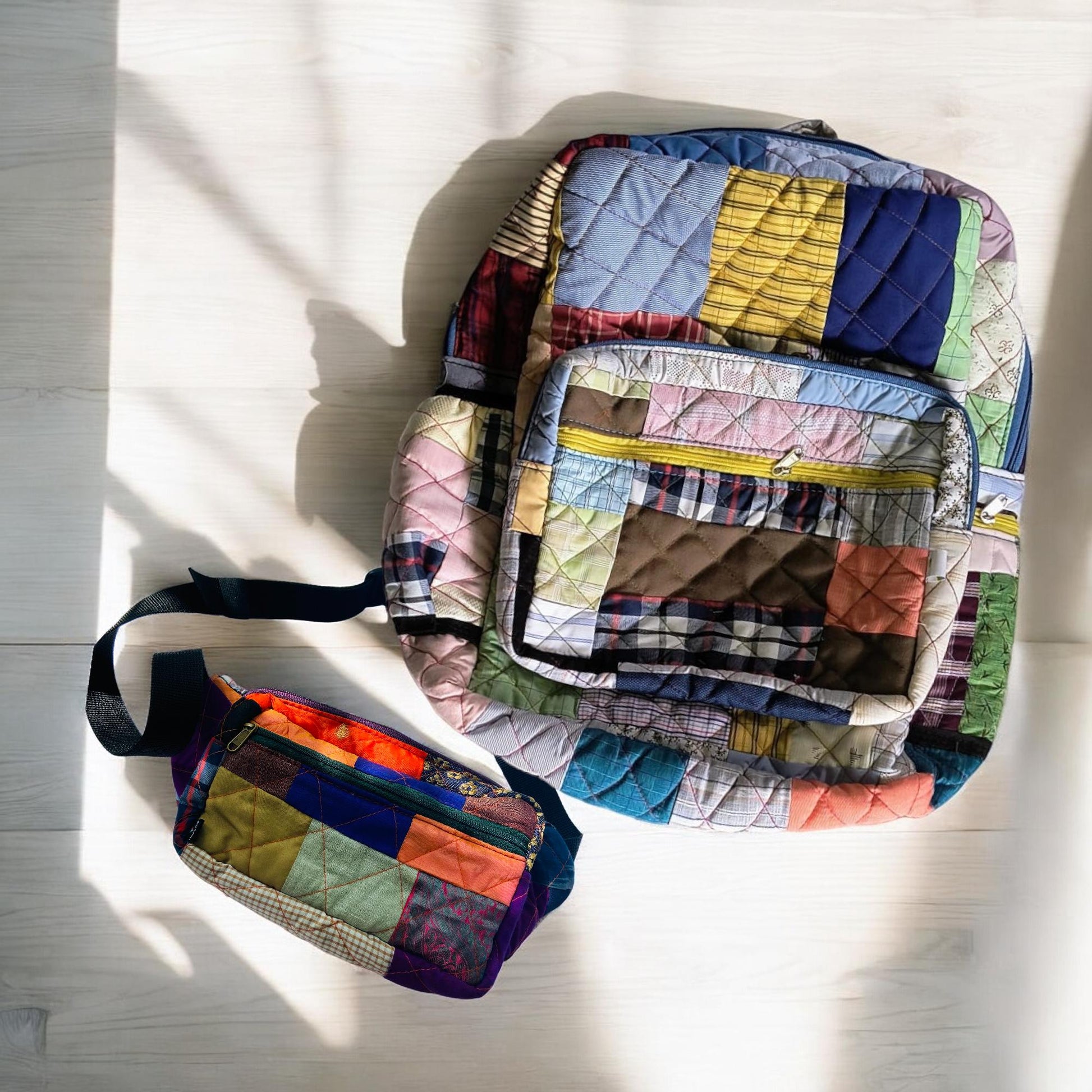 Quilted backpack and fanny pack made from colorful patchwork laid on a light wooden floor. Handmade using fabric scraps, these bags are ideal for travel, hikes, or school use. The fanny pack has a mix of bold colors including purple, orange, and green, with an adjustable strap. Sustainable, practical, and stylish, perfect for eco warriors and lovers of upcycled, one-of-a-kind accessories.