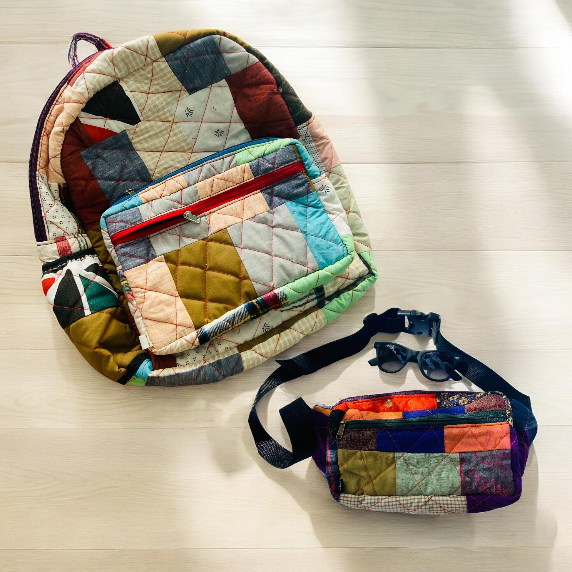 Patchwork backpack and matching fanny pack shown on a light wooden floor with sunglasses placed nearby. Each bag is made from vibrant upcycled cotton fabrics and finished with sturdy zippers. A perfect travel set for road trips, beach days, or music festivals. Ideal for those who value handmade, zero-waste, colorful accessories that blend style and sustainability effortlessly.