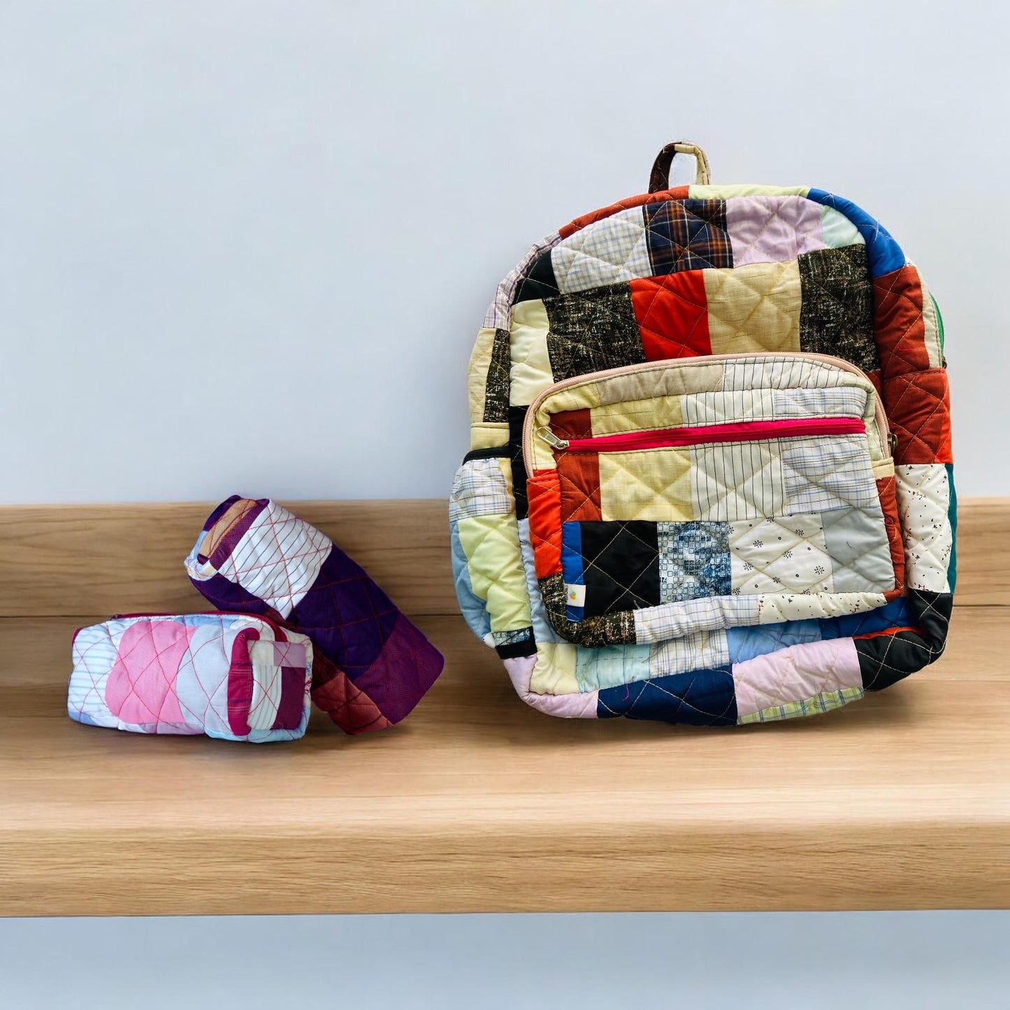 Backpack and two pouches displayed on a wooden shelf. The backpack features multicolored patchwork quilting with a front red zipper pocket, paired with matching eco-friendly pouches. Stylish, handcrafted, and sustainable โ perfect for travel, school, beach, or gifting. Great for toddlers, women, or anyone looking for handmade, upcycled backpacks and organizational sets with a unique patchwork aesthetic.