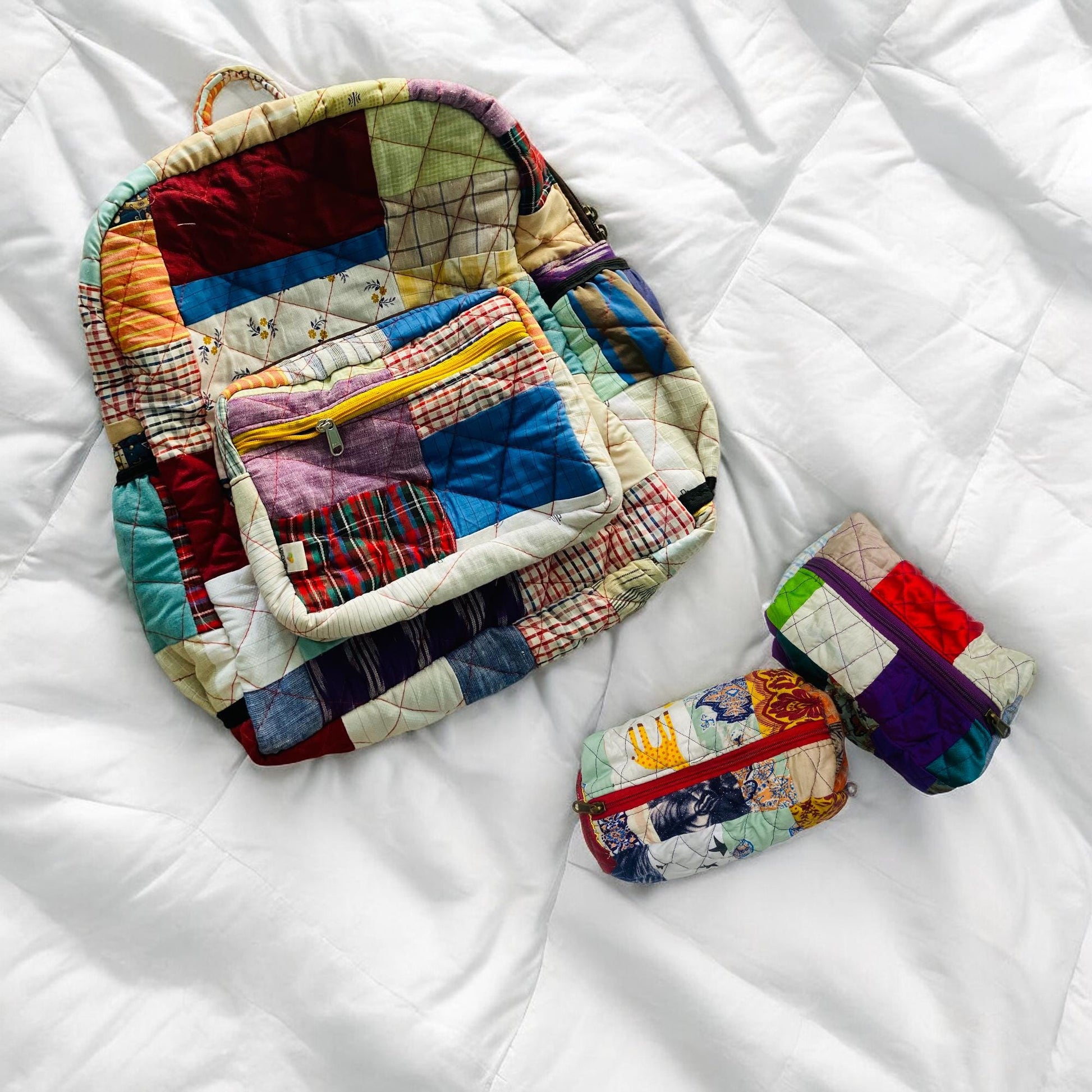 Colorful backpack and quilted pouches resting on a white duvet. Made using patchwork of various upcycled fabrics and vibrant zippers, the set is a cheerful, sustainable alternative to plastic bags. Great for school, travel, or gifting. Handcrafted by artisans, it suits toddlers, teens, and women. Perfect for baby shower gifts or as a unique accessory set for minimalist and conscious living.