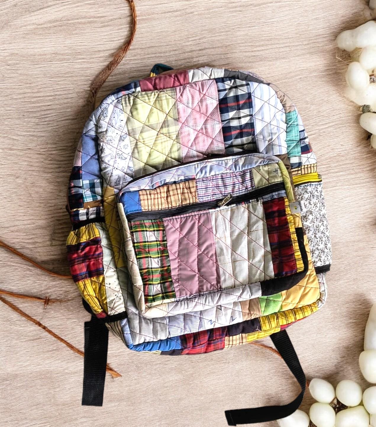 Single handcrafted quilted patchwork backpack in pastel tones placed on a wooden surface. The bag features neat diamond stitching, multiple compartments, and adjustable black straps. Ethically made using rescued tailoring waste. Ideal for travel, school, or gifting, this unique backpack combines durability, comfort, and sustainability with a colorful and artistic twist.