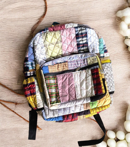 Single handcrafted quilted patchwork backpack in pastel tones placed on a wooden surface. The bag features neat diamond stitching, multiple compartments, and adjustable black straps. Ethically made using rescued tailoring waste. Ideal for travel, school, or gifting, this unique backpack combines durability, comfort, and sustainability with a colorful and artistic twist.