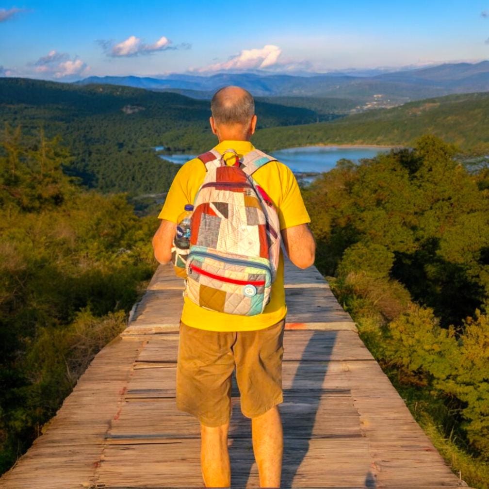 Back view of a man wearing a sustainable patchwork backpack while overlooking a forested mountain landscape. The handmade bag is made from recycled cotton fabrics in earthy tones, showcasing its functionality and comfort for outdoor adventures. Perfect for hiking, travel, or daily use. Ideal for eco-conscious explorers who appreciate both nature and sustainable craftsmanship.