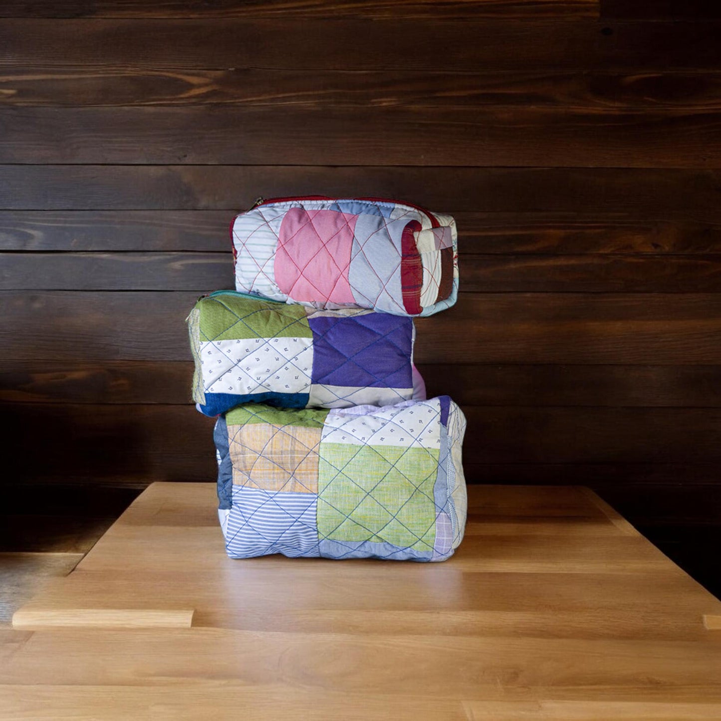 Set of three handmade quilted fabric pouches stacked on a wooden surface against a dark wood wall. Each pouch is made from colorful upcycled fabric squares stitched together. These eco-friendly travel organizers are ideal for makeup, toiletries, school supplies, or gifting. A perfect sustainable gift for eco warriors, students, or minimalist travelers who care about zero-waste lifestyles and handmade storage.