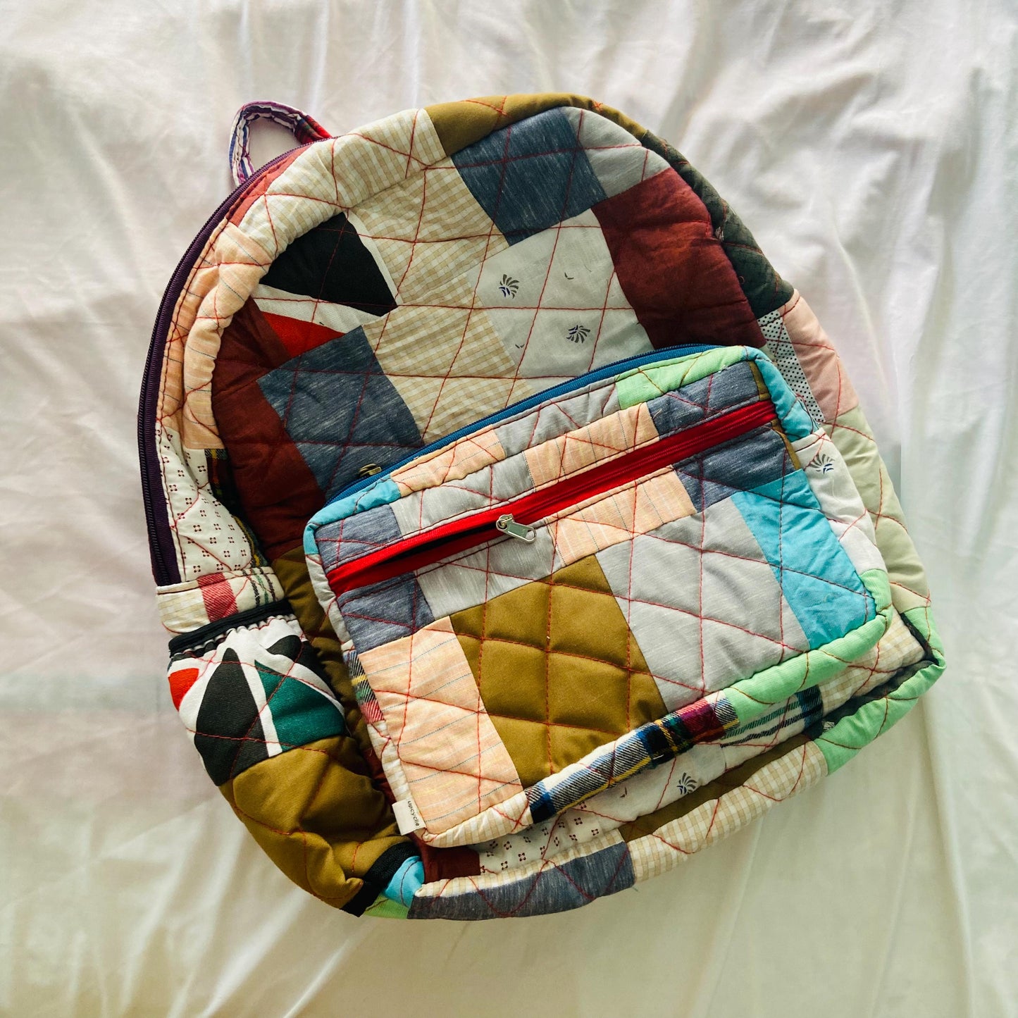 Boho-style patchwork backpack made from rescued cotton textiles, displayed on a white fabric background. Featuring earthy tones, red and pink accents, and a bold magenta zipper, this backpack is a practical and aesthetic choice for school, travel, or gifting. Handmade, quilted, and eco-conscious, it promotes a zero-waste lifestyle through its functional, upcycled design.