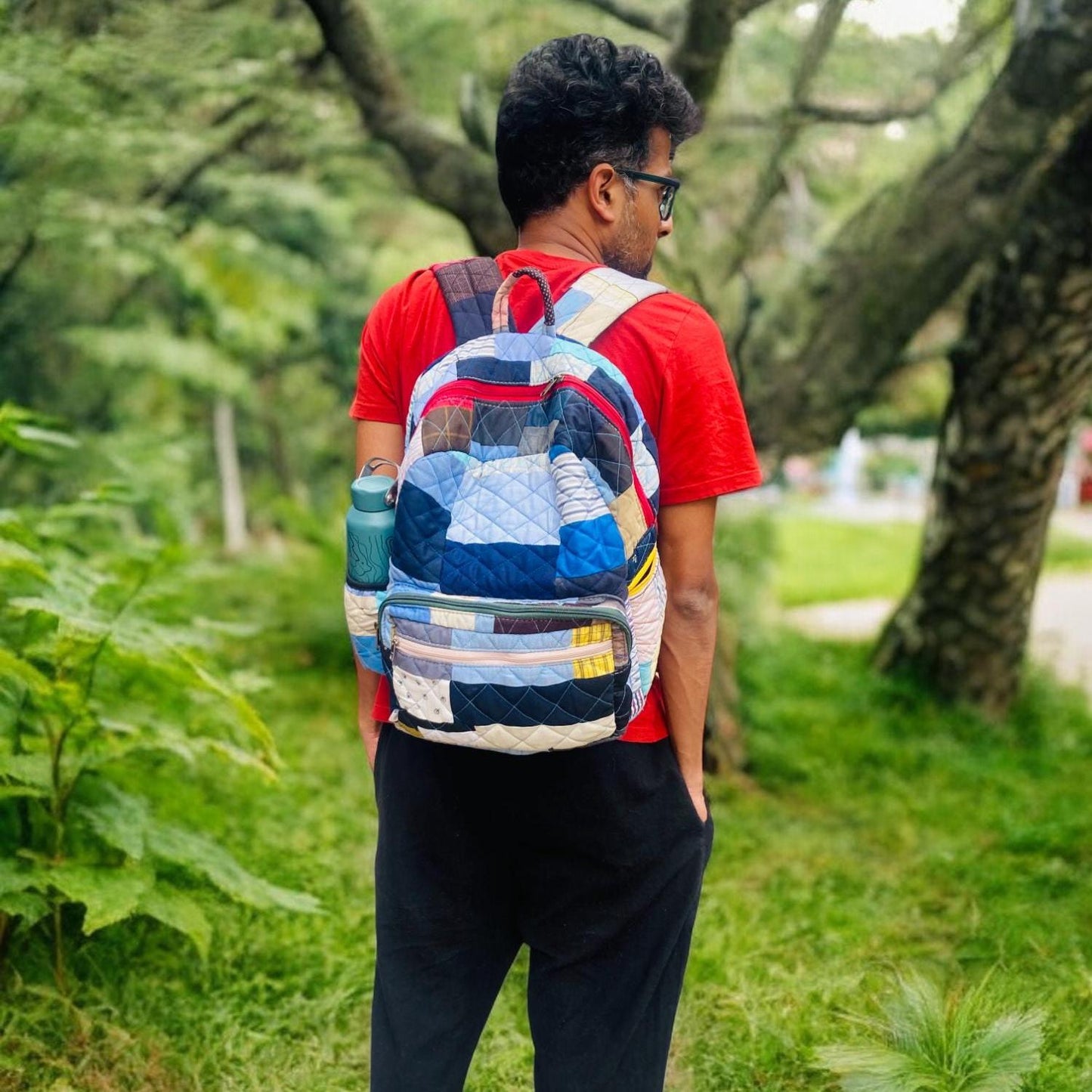 Eco-conscious man wearing a colorful patchwork backpack while standing in a lush green forest. The bag, made from recycled cotton fabrics, includes a side pocket with a water bottle. Designed for functionality and flair, it’s perfect for trekking, school, or city use. A durable and vibrant choice for ethical consumers who enjoy outdoor exploration and handcrafted style.