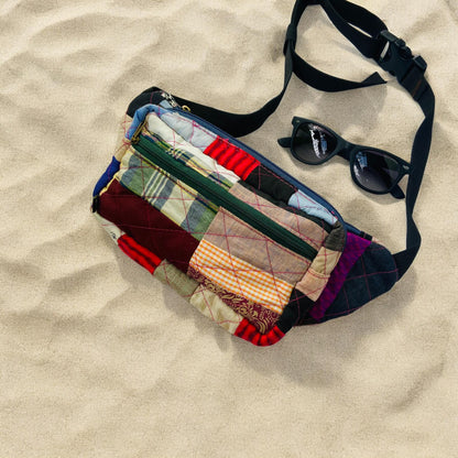Eco-friendly patchwork fanny pack lying on sandy beach next to sunglasses. Designed for travel and hands-free convenience, this handmade piece showcases bold fabric combinations in red, green, beige, and purple. Features durable zippers and an adjustable black strap. Perfect for beach days, hikes, or urban adventures. A zero-waste accessory for those who love function with fashion.